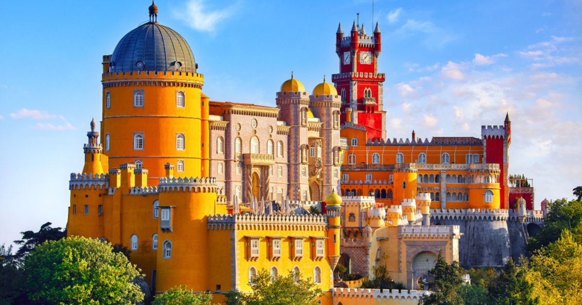 The colorful Palace of Pena in Sintra, outside of Lisbon. The building is a mix of deep golden yellow, red, orange, and brown walls, curved and straight lines, and towers. Below are green-leaved trees, and the sky is blue with thin white clouds overhead.