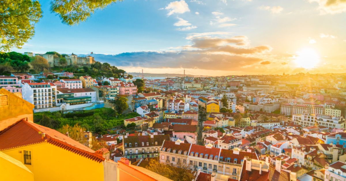 Panoramic view of Lisbon at sunset, with red roofs and colorful houses and buildings stretching out into the distance, the corner of a golden yellow building in the foreground, and light blue sky with thick clouds overhead.