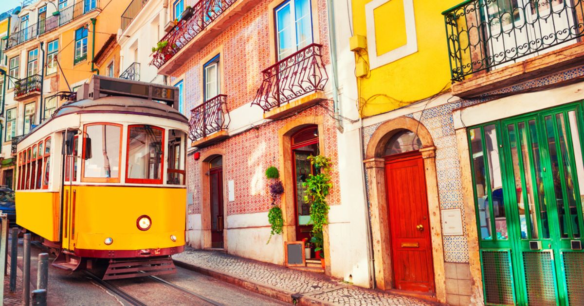 Vintage yellow tram on a narrow street in Lisbon, with a row of bright, colorful buildings and stone sidewalk alongside it.