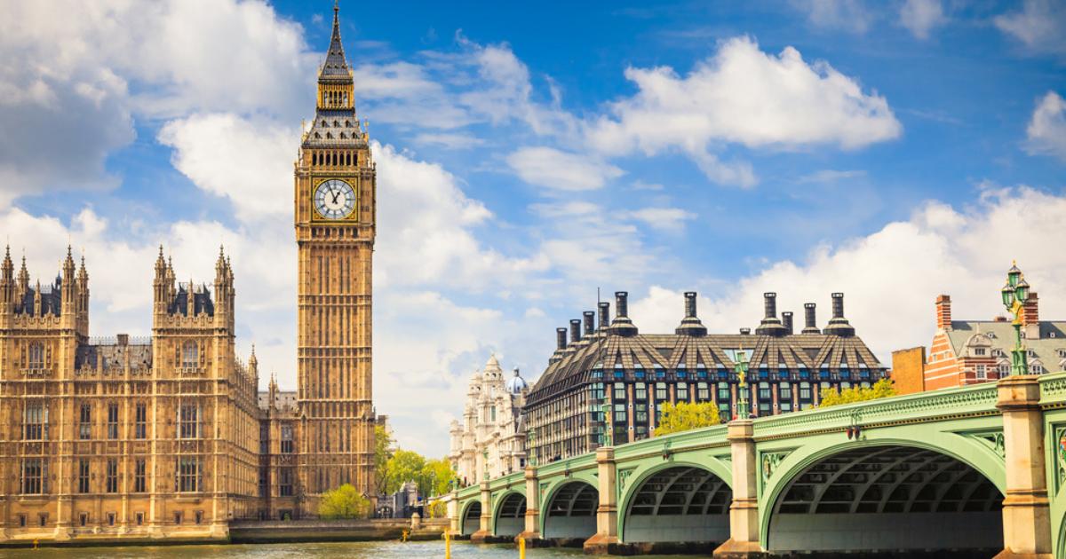 Big Ben, Houses of Parliament, a bridge, and other buildings, alongside the Thames River with blue sky and white, fluffy clouds overhead.