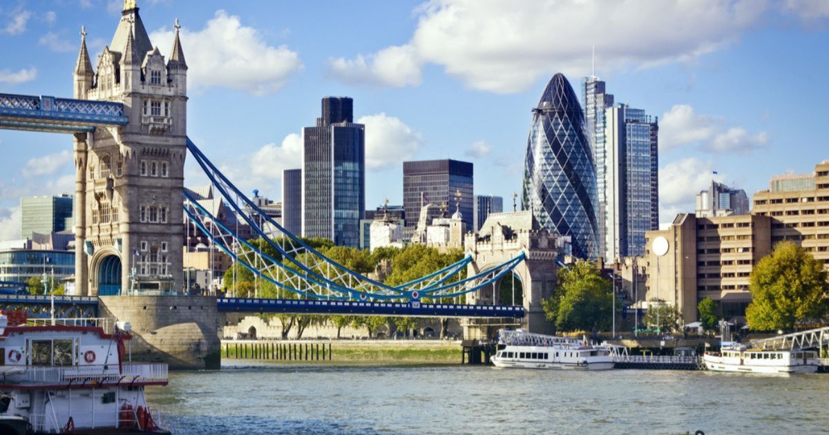 Wide shot of the Tower Bridge over the Thames, with the financial district buildings in the background, large boats in the water, and light blue sky with white clouds overhead.