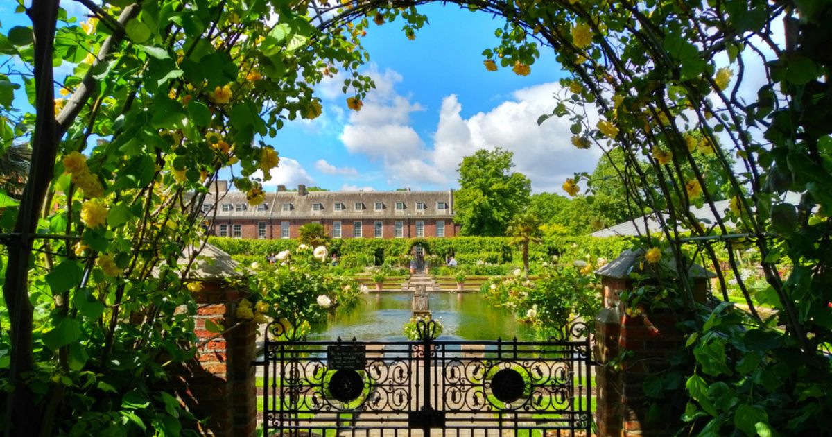 View of Kensington Gardens through a shaded archway of blooming yellow flowers and green leaves, with garden gate, pond, green hedges, and a brick building in the background. The sky is blue with white clouds.