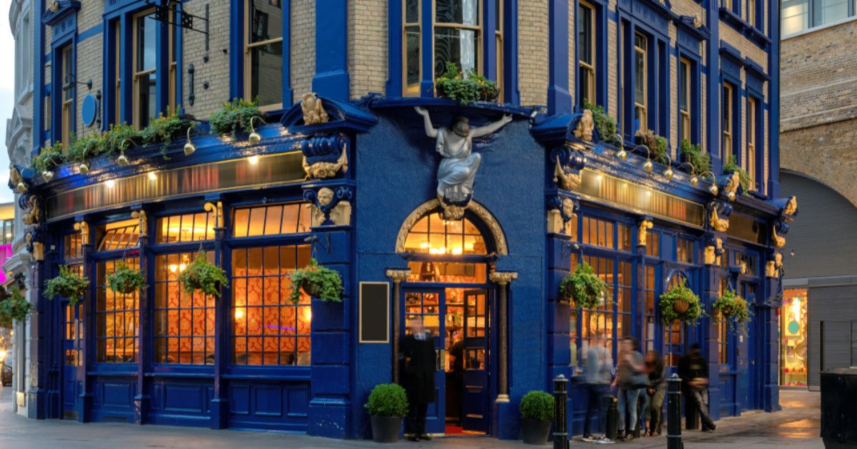 Twilight photo of a traditional central London pub. The building is light brown brick and dark blue walls, with large windows lit with golden light from inside on all sides, an arched doorway, and green plants hanging in baskets outside.